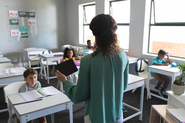 Junge weiße Lehrerin mit digitalem Tablet spricht mit multiracialen Grundschülern