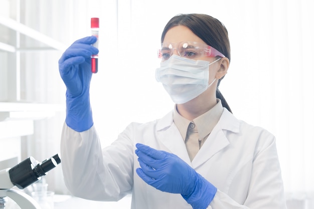 Junge Wissenschaftlerin oder Chemikerin, die Kolben mit flüssiger Substanz hält, während Eigenschaften des neuen Impfstoffs im Labor untersucht