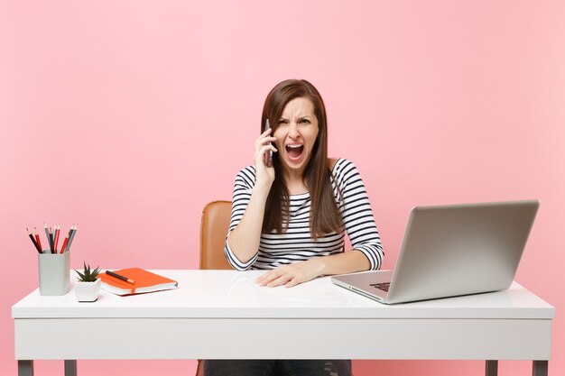 Junge wütende Frau, die schreit und mit dem Handy spricht, während sie mit PC-Laptop im Büro sitzt und an einem Projekt arbeitet?