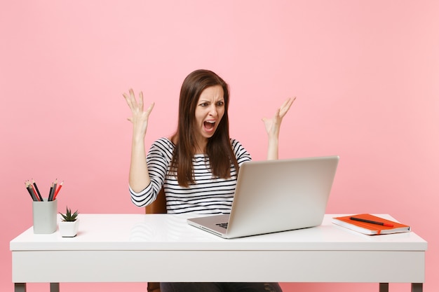 Junge wütende Frau in Freizeitkleidung, die schreit, hat Probleme, Hände zu verteilen, die mit Laptop am Projekt arbeiten, während sie im Büro sitzen