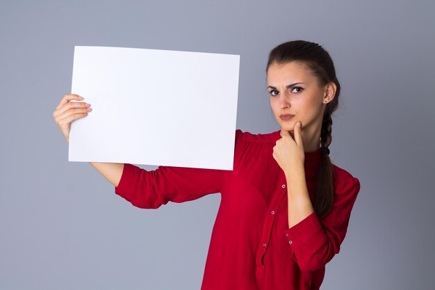 Junge wundernde Frau in roter Bluse mit weißem Blatt Papier auf grauem Hintergrund im Studio