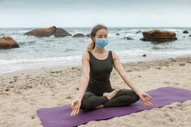 Junge Yogi-Frau in Sportuniform meditiert in einer medizinischen Maske, die auf einer Matte an den Stränden sitzt Harmonie