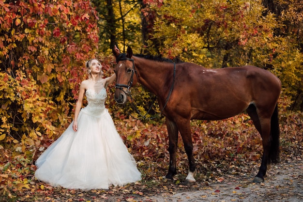 Junge zarte Braut in einem üppigen weißen Kleid posiert in einem Herbstpark mit einem Pferd