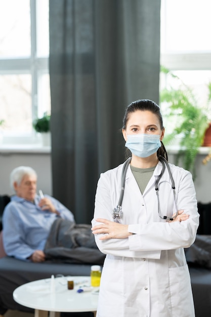 Junge zeitgenössische Ärztin in Weißkittel und Schutzmaske, die ihre Arme an der Brust verschränkt und Sie gegen einen kranken älteren Patienten im Bett ansieht