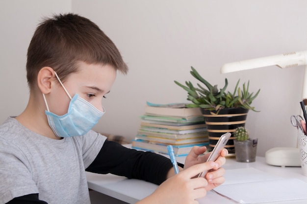 Junge zu Hause, der mit einem Smartphone in seinen Händen und einer medizinischen Maske schult. Coronavirus-Quarantänekonzept