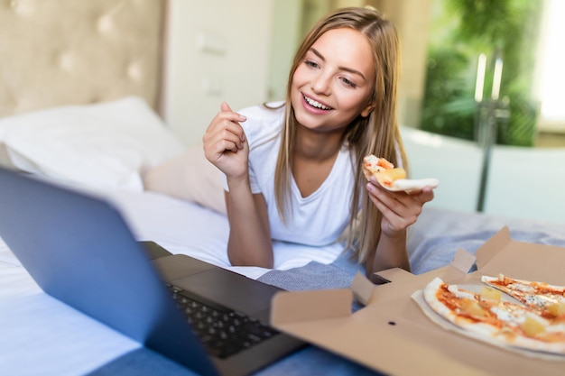 Junge zufällige Frau, die zu Hause auf einem Bett im Bett liegt, während sie Pizza isst und einen Laptop benutzt?