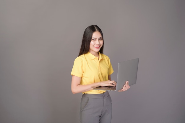 Junge zuversichtliche schöne Frau, die gelbes Hemd trägt, hält einen Laptop auf grauem Hintergrundstudio