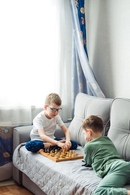 Jungen, die Holzschach spielen, liegen auf einem grauen Sofa vor dem Fenster