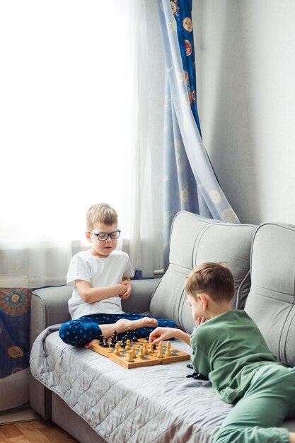 Jungen, die Holzschach spielen, liegen auf einem grauen Sofa vor dem Fenster