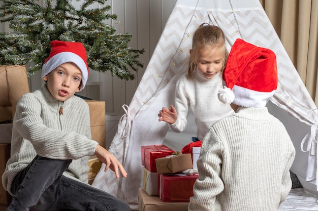 Jungen in einer Weihnachtsmütze geben einem Mädchen zu Weihnachten und Neujahr im Kindergarten Geschenke. Brüder und Schwestern tauschen Geschenke aus