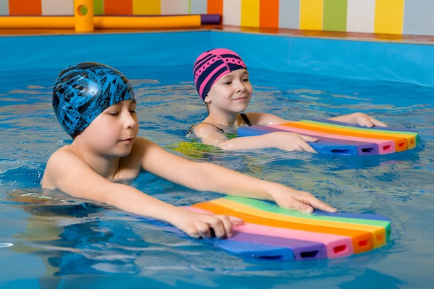 Jungen und Mädchen im Badeanzug üben mit einem Schaumstoffpolster das Schwimmen im Schwimmbad