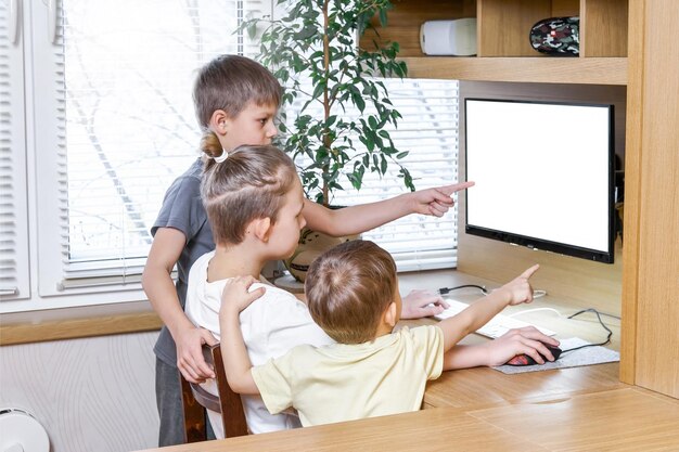 Jungen zeigen einen leeren weißen Computerbildschirm, der mit Fingern zeigt, die auf einem Holzschreibtisch sitzen und stehen