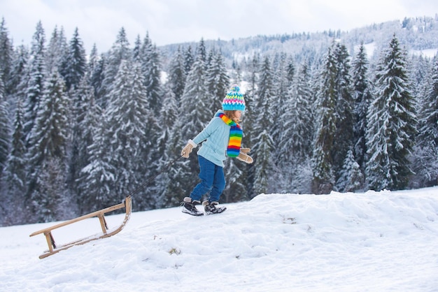 Jungenkind, das mit Schlitten in den Winterschnee-Weihnachtskinderfeiertagen und guten Rutsch ins Neue Jahr-Schneedecke schiebt