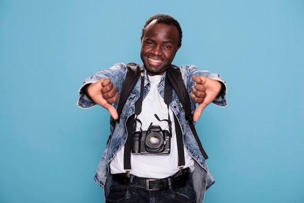 Junger afrikanisch-amerikanischer Fotografie-Enthusiast zeigt missbilligende Handgesten, während er auf blauem Hintergrund steht. Lächelnder Fotograf, der das negative Symbol mit dem Daumen nach unten gibt, während er eine DSLR-Kamera hat.