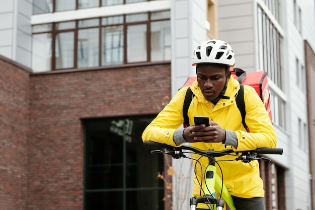 Junger afrikanisch-amerikanischer Lieferbote mit Handy auf Fahrrad sitzend