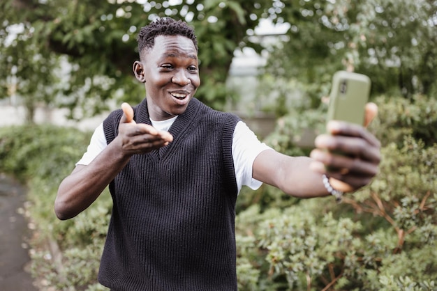 Junger afrikanischer Mann, der Selfie oder Vlogging am Telefon im Park macht und lächelt