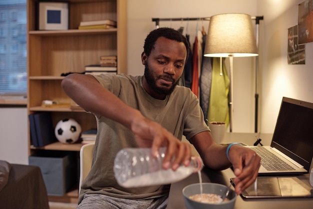 Junger afrikanischer Mann gießt Milch in eine Schüssel mit Müsli, während er morgens am Arbeitsplatz frühstückt