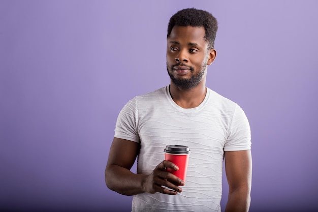 Junger afrikanischer mann mit kurzen lockigen haaren, der seine freizeit genießt, ein lässiges weißes t-shirt trägt, kaffee zum mitnehmen hält, einzeln auf violettem hintergrund, leckeres getränk trinkt. Menschen und Freizeit.