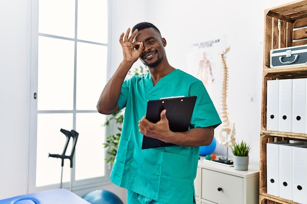 Junger afrikanischer Physiotherapeut, der in der Klinik für Schmerztherapie arbeitet, lächelt glücklich und macht ein „OK“-Zeichen mit der Hand auf dem Auge, die durch die Finger schaut
