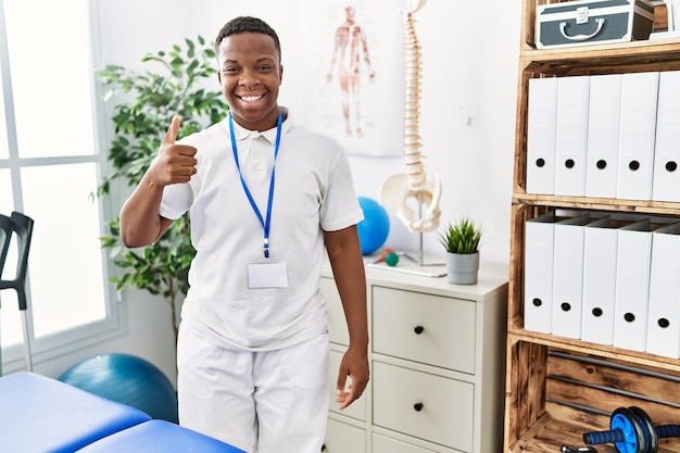 Junger afrikanischer physiotherapeut, der in einer schmerzheilungsklinik arbeitet, lächelt glücklich und positiv, daumen hoch, tut ausgezeichnet und genehmigungszeichen