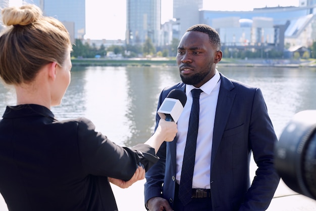 Junger afrikanischer Unternehmer, der während des Interviews im städtischen Umfeld vor einer Reporterin oder Journalistin mit Mikrofon steht