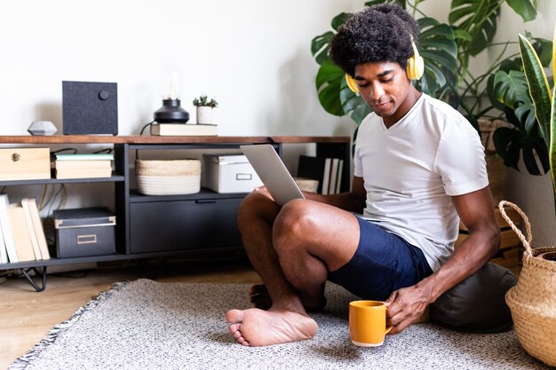 Junger Afroamerikaner, der Laptop und Kopfhörer benutzt und Morgenkaffee in seinem hellen Wohnzimmer trinkt. Platz kopieren. Konzept zu Hause. Technologiekonzept.