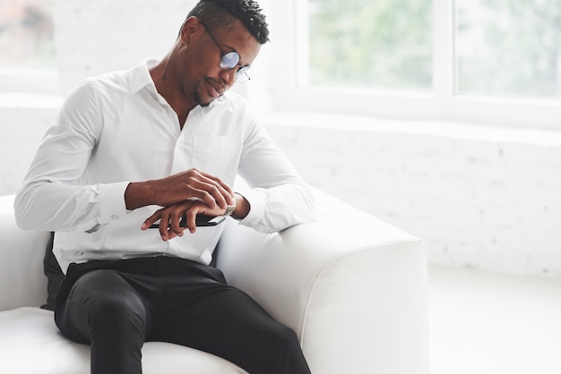 Junger Afroamerikaner in Bürokleidung sitzt auf dem weißen Sofa