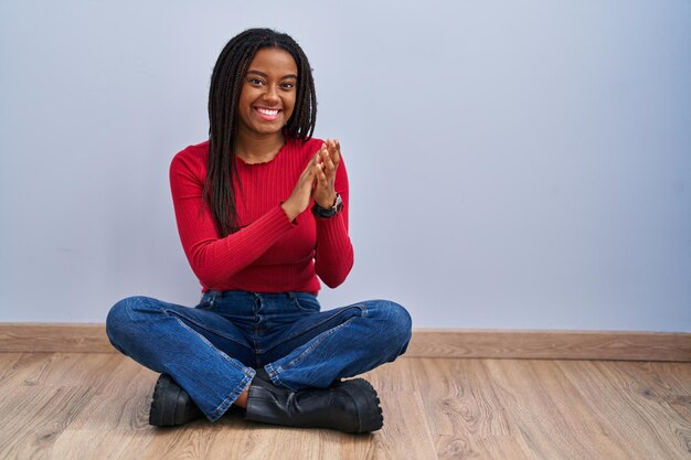 Junger Afroamerikaner mit Zöpfen, der zu Hause auf dem Boden sitzt, klatscht und applaudiert glücklich und fröhlich, lächelnde stolze Hände zusammen