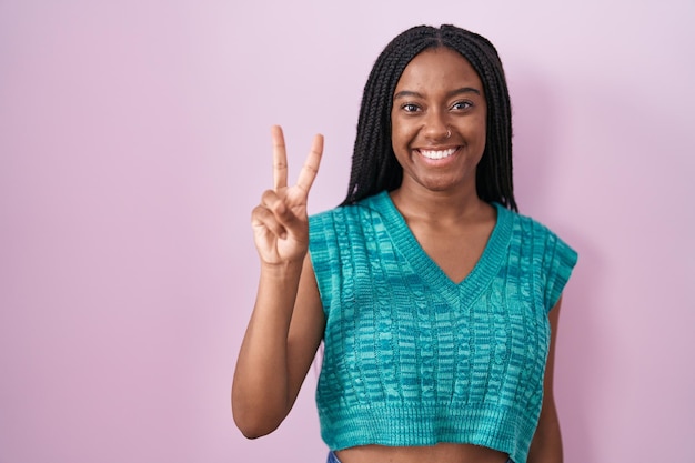 Junger Afroamerikaner mit Zöpfen steht vor rosa Hintergrund und zeigt mit den Fingern Nummer zwei nach oben, während er selbstbewusst und glücklich lächelt