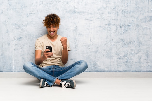 Junger Afroamerikanermann, der auf dem Boden mit Telefon in Siegposition sitzt