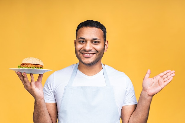 Junger afroamerikanischer indischer schwarzer Mann isst Hamburger isoliert über gelbem Hintergrund. Kochen Sie, das Burger zubereitet.