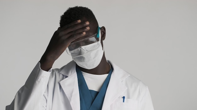 Junger afroamerikanischer männlicher Arzt mit Schutzbrille und medizinischer Maske posiert auf grauem Hintergrund Müde nach der Arbeit Ausdruck