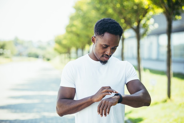 Junger afroamerikanischer Mann, bei einem morgendlichen Joggen in einem weißen T-Shirt im Park, wechselt das Sportprogramm in einem Smartwatch-Fitnessarmband, das sich mit einem Fitness-aktiven Lebensstil beschäftigt