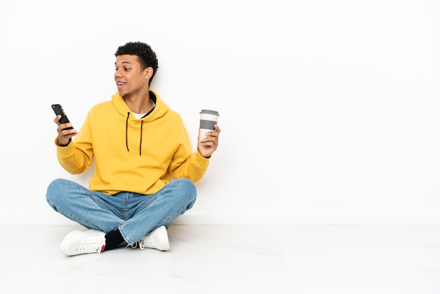 Junger afroamerikanischer Mann, der auf dem Boden sitzt, isoliert auf weißem Hintergrund, der Kaffee zum Mitnehmen und ein Handy hält