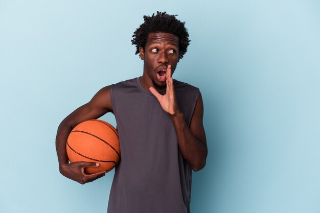 Junger afroamerikanischer Mann, der Basketball isoliert auf blauem Hintergrund spielt, sagt eine geheime heiße Bremsnachricht und schaut beiseite