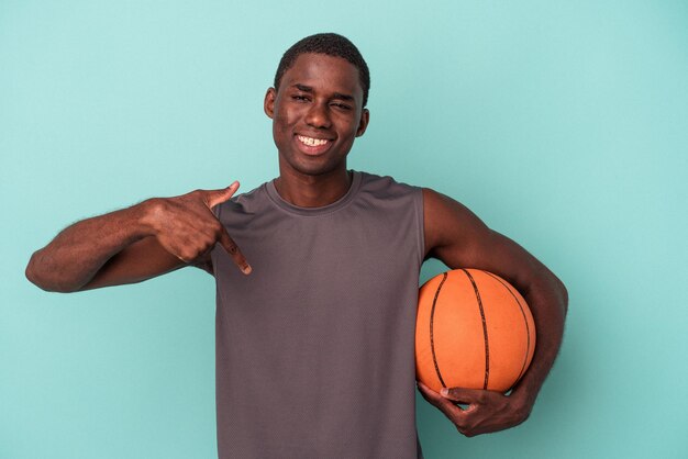 Junger afroamerikanischer Mann, der Basketball spielt, isoliert auf blauem Hintergrund, zeigt mit der Hand auf einen Hemdkopienraum, stolz und selbstbewusst