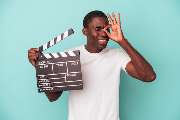Junger afroamerikanischer Mann, der die Klappe isoliert auf blauem Hintergrund hält, aufgeregt, die OK-Geste im Auge zu behalten