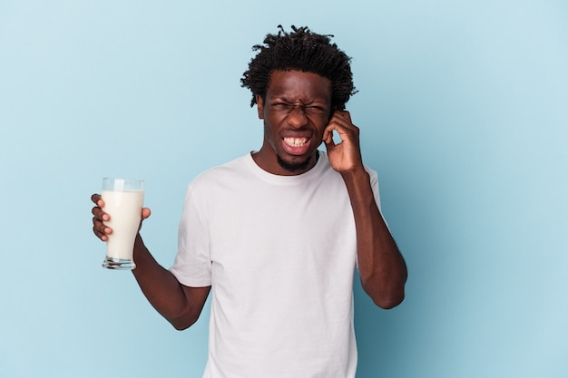 Junger afroamerikanischer Mann, der ein Glas Milch auf blauem Hintergrund isoliert hält und die Ohren mit den Händen bedeckt.