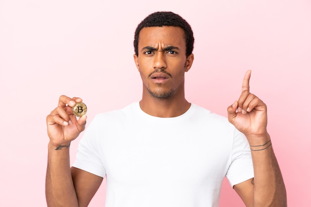 Junger afroamerikanischer Mann, der einen Bitcoin über isoliertem rosa Hintergrund hält und eine Idee denkt, die mit dem Finger nach oben zeigt