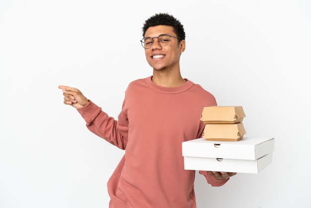 Junger afroamerikanischer Mann, der einen Burger und Pizzas auf weißem Hintergrund hält, der mit dem Finger zur Seite zeigt