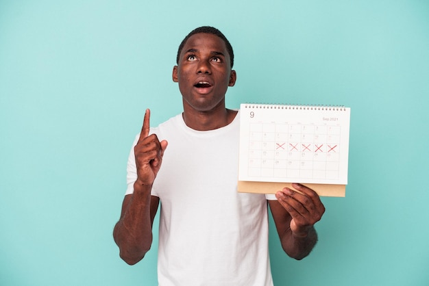 Junger afroamerikanischer Mann, der einen Kalender isoliert auf blauem Hintergrund hält und mit geöffnetem Mund nach oben zeigt.