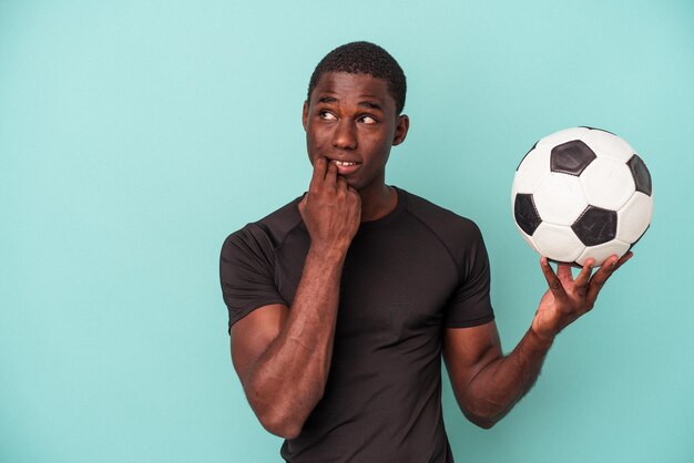 Junger afroamerikanischer Mann, der Fußball spielt, isoliert auf blauem Hintergrund, dachte entspannt über etwas nach und betrachtete einen Kopierraum.