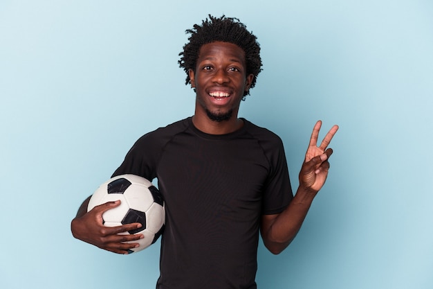 Junger afroamerikanischer Mann, der Fußball spielt, isoliert auf blauem Hintergrund, fröhlich und sorglos, der ein Friedenssymbol mit den Fingern zeigt.