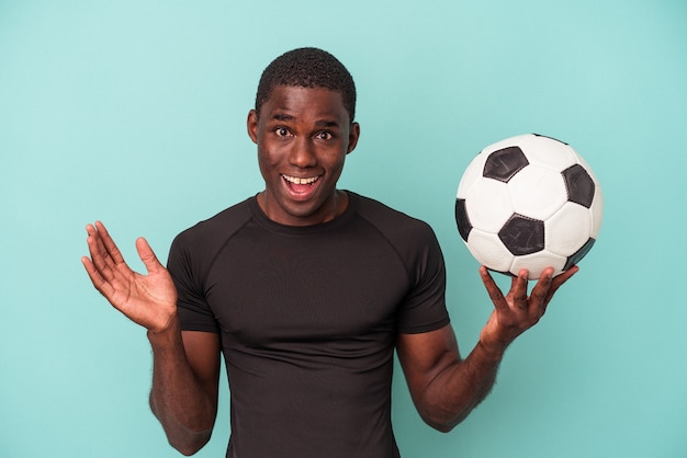 Junger afroamerikanischer Mann, der Fußball spielt, isoliert auf blauem Hintergrund, überrascht und schockiert.