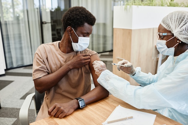 Junger afroamerikanischer Mann, der in der Arztpraxis sitzt und sich eine Grippeimpfung in den Arm bekommt?