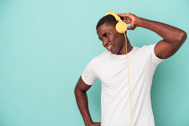 Junger afroamerikanischer Mann, der Musik hört, isoliert auf blauem Hintergrund, der den Hinterkopf berührt, denkt und eine Wahl trifft.