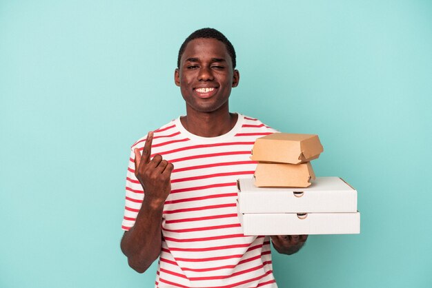 Junger afroamerikanischer Mann, der Pizza und Burger einzeln auf blauem Hintergrund hält und mit dem Finger auf Sie zeigt, als ob er einladen würde, näher zu kommen.