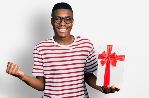 Junger afroamerikanischer Mann hält ein Geschenk in der Hand und schreit stolz, feiert Sieg und Erfolg, sehr aufgeregt mit erhobenem Arm