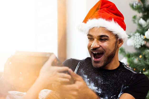 Junger afroamerikanischer Mann in Weihnachtsmütze freut sich, sein Weihnachtsgeschenk zu erhalten