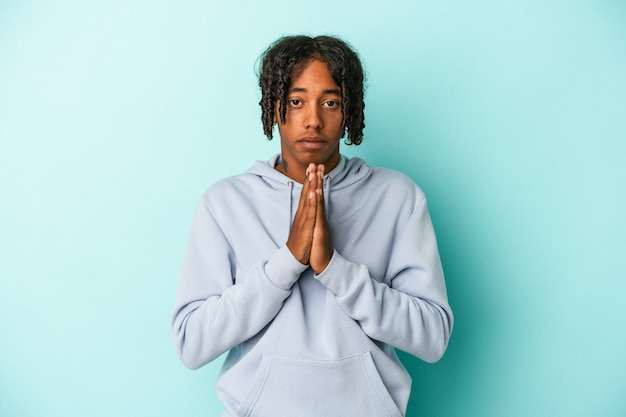 Junger afroamerikanischer Mann isoliert auf blauem Hintergrund betend, Hingabe zeigend, religiöse Person, die nach göttlicher Inspiration sucht.